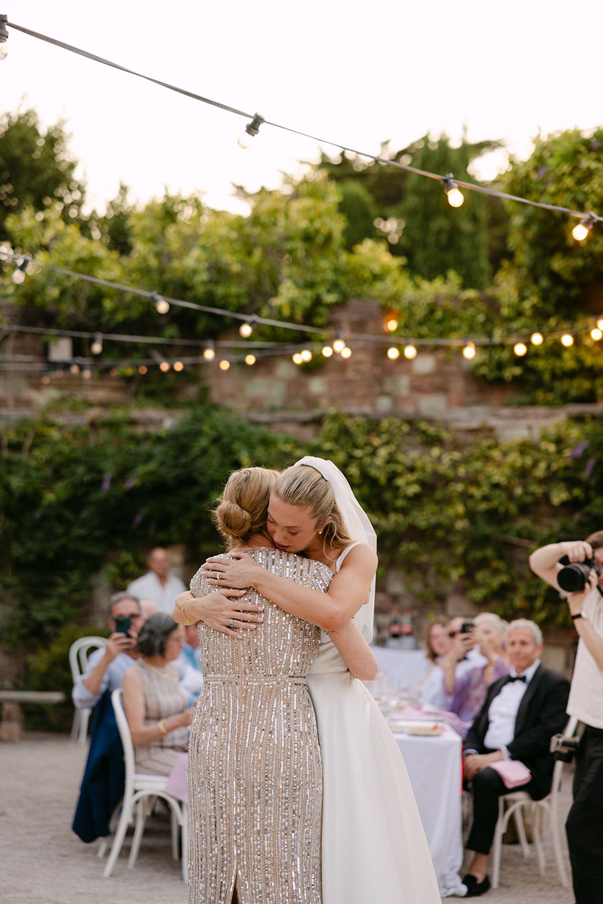 The bride embraces a guest in a sequinned gown in a lantern-lit courtyard while seated guests look on.