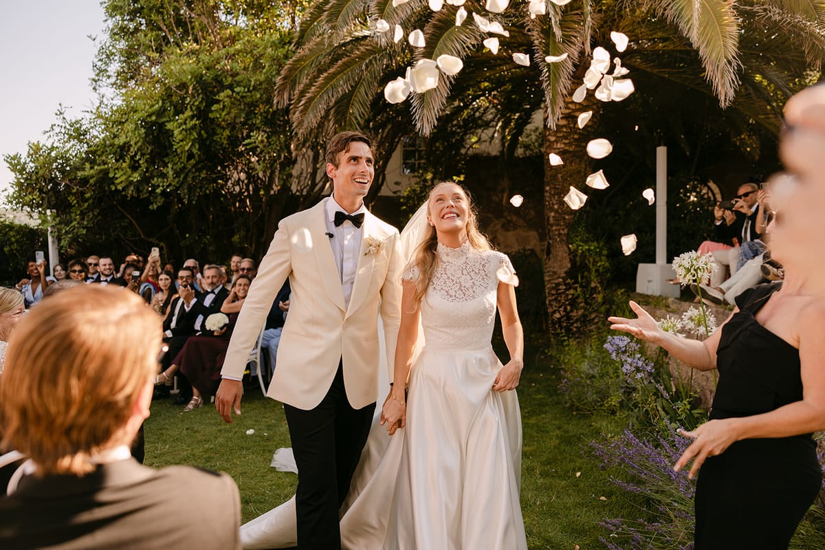 The couple walk hand in hand across the lawn as guests toss white petals into the air around them.