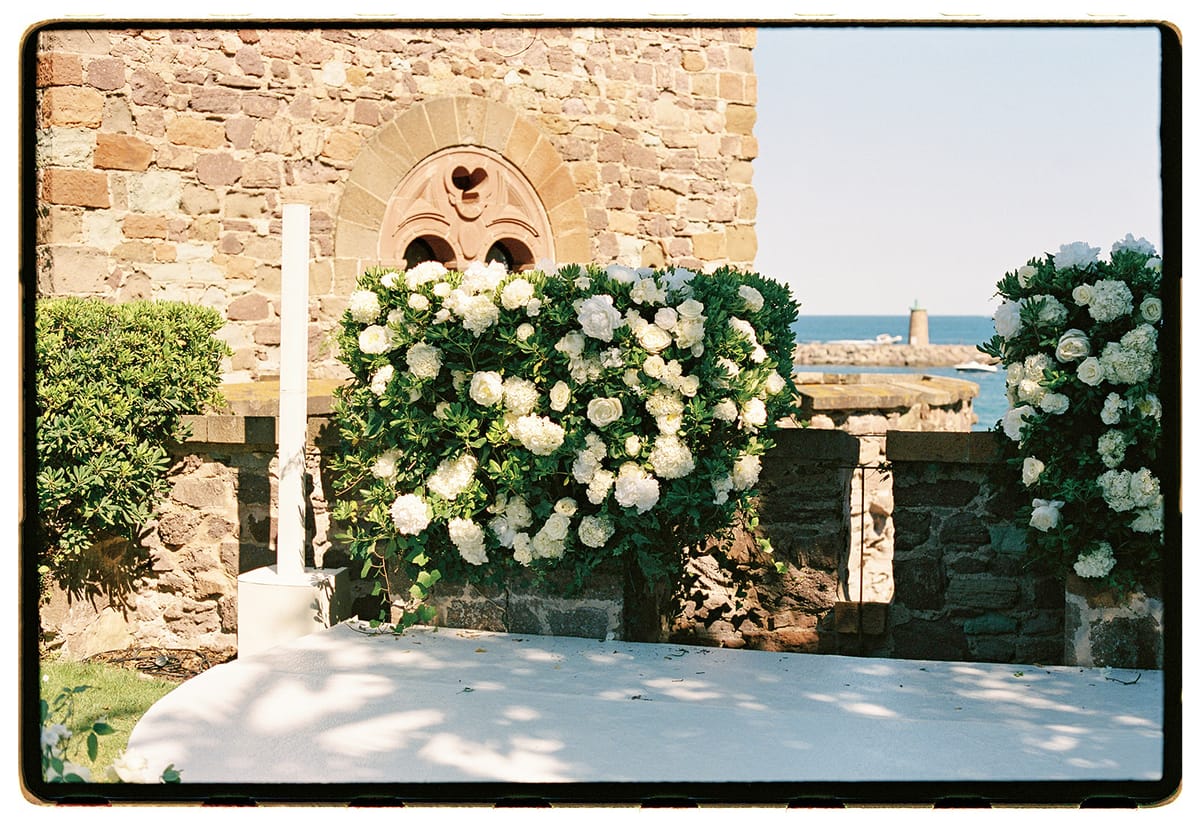 Large white hydrangea arrangements and a tall candle sit on a white platform beside a stone wall, with the sea behind.