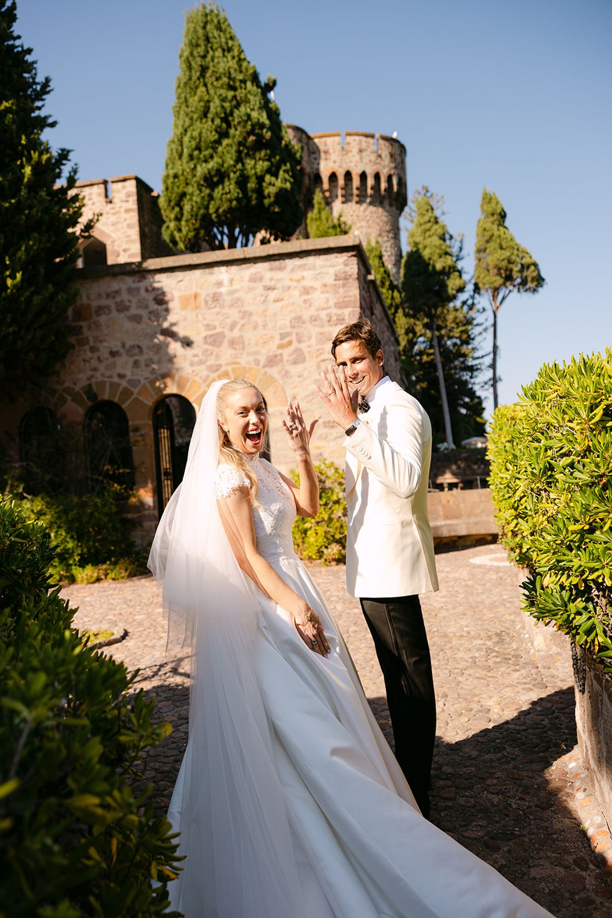 The bride in a long veil and the groom in a white jacket smile at the camera in a garden beside a stone castle.