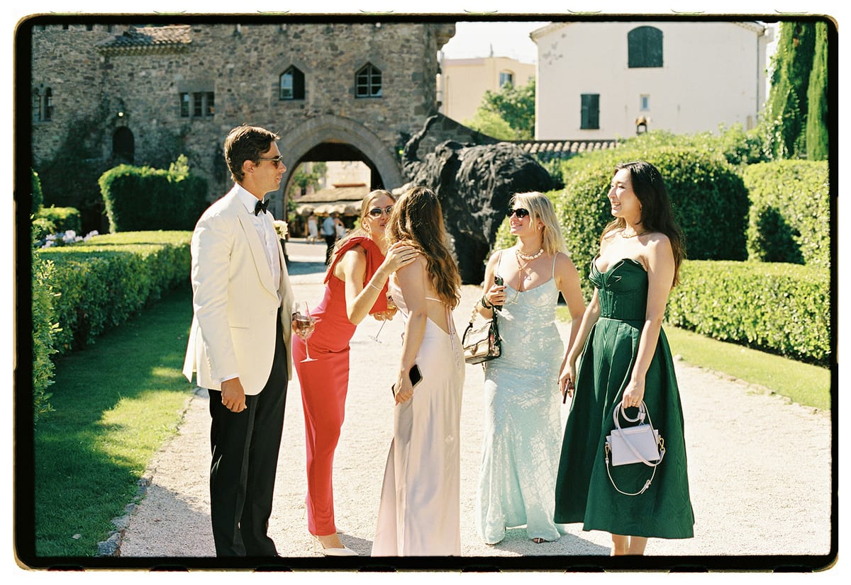The groom in a white jacket and sunglasses talks with four women in colourful dresses on a gravel path beside clipped hedges.
