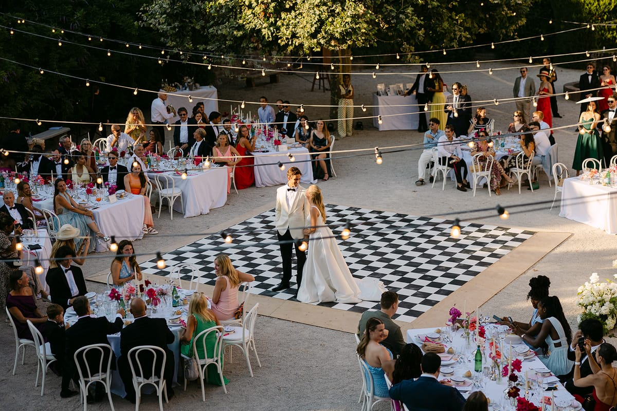 The couple dance on a black and white chequered floor in a courtyard, surrounded by guests at round tables under festoon lights.