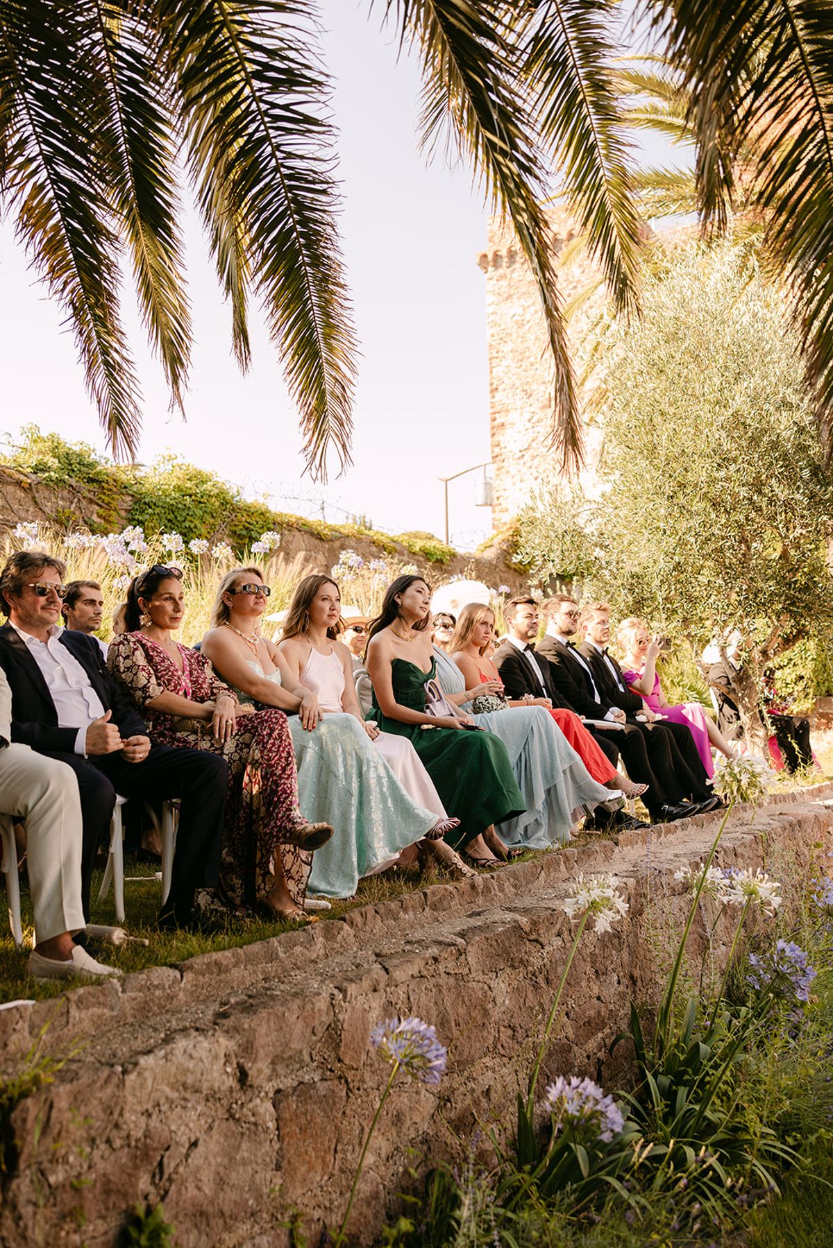 Guests in colourful outfits sit in a line along an old stone wall beneath palm fronds in warm sunlight.