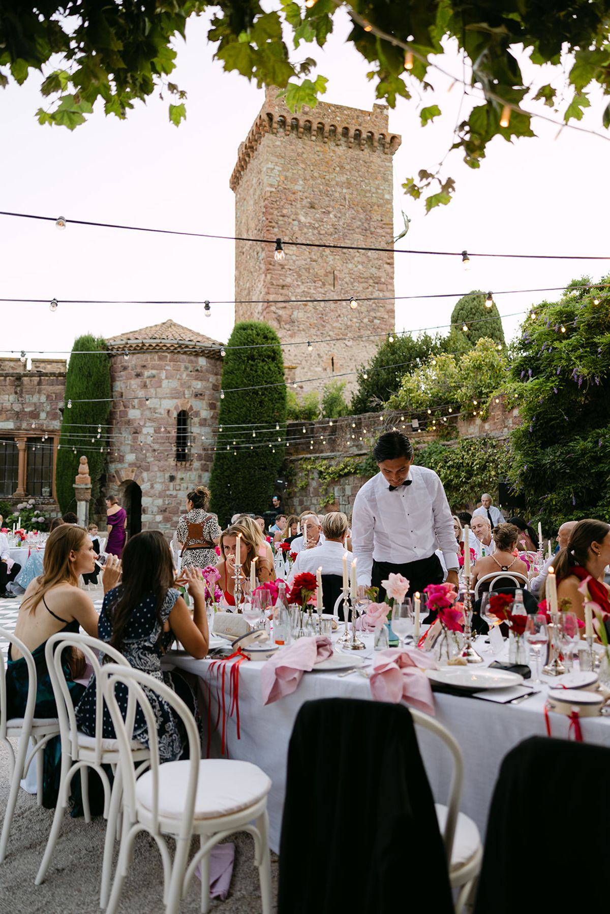 Guests sit at round tables with pink flowers and ribbons beneath string lights, with a stone tower rising behind.