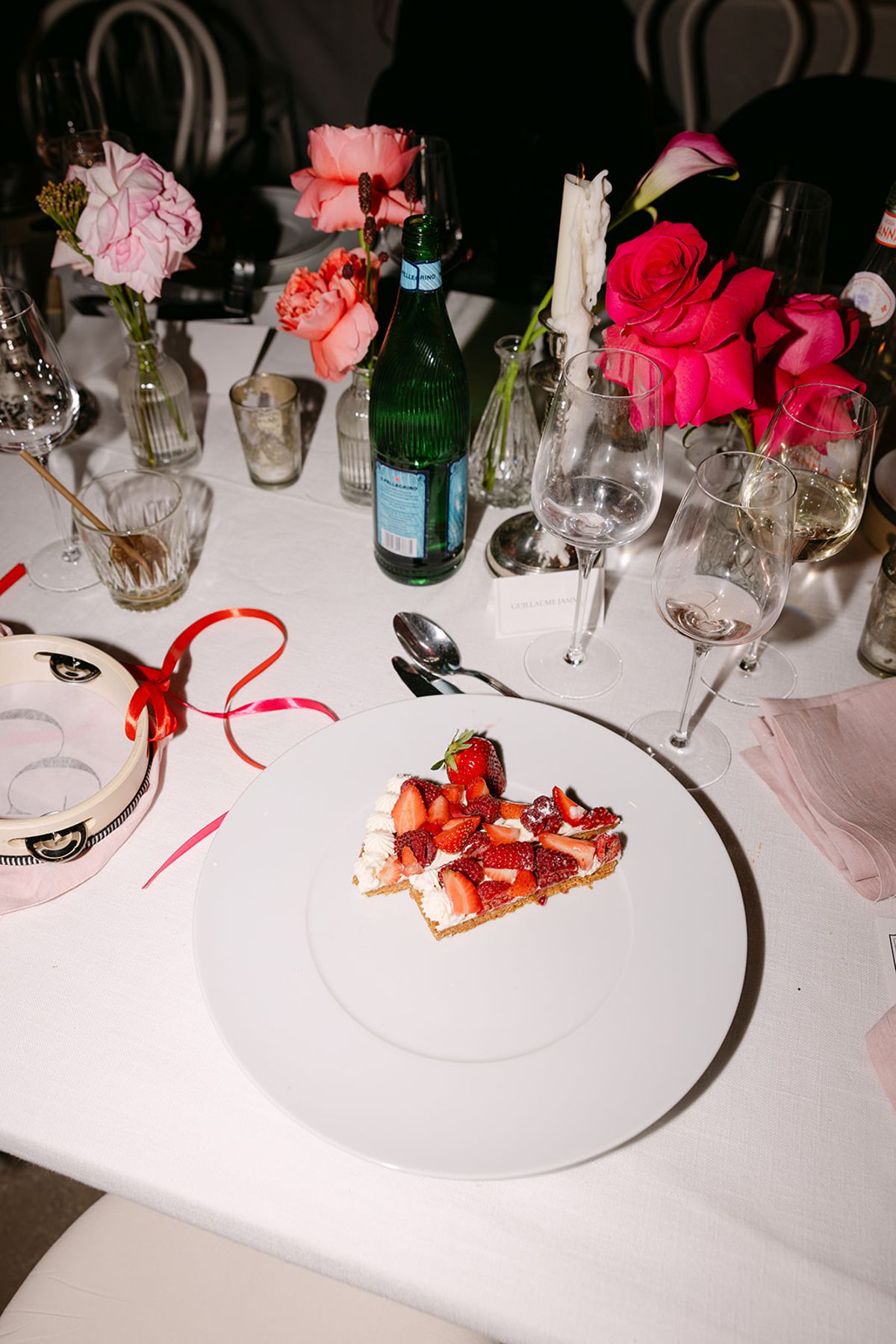 A white-clothed table set with pink and red flowers, glasses, a green bottle and a plate of strawberry dessert.