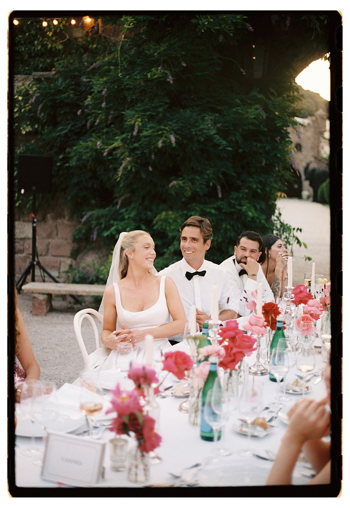 The bride and groom sit at a dinner table with a third person, smiling, beside vases of pink flowers.