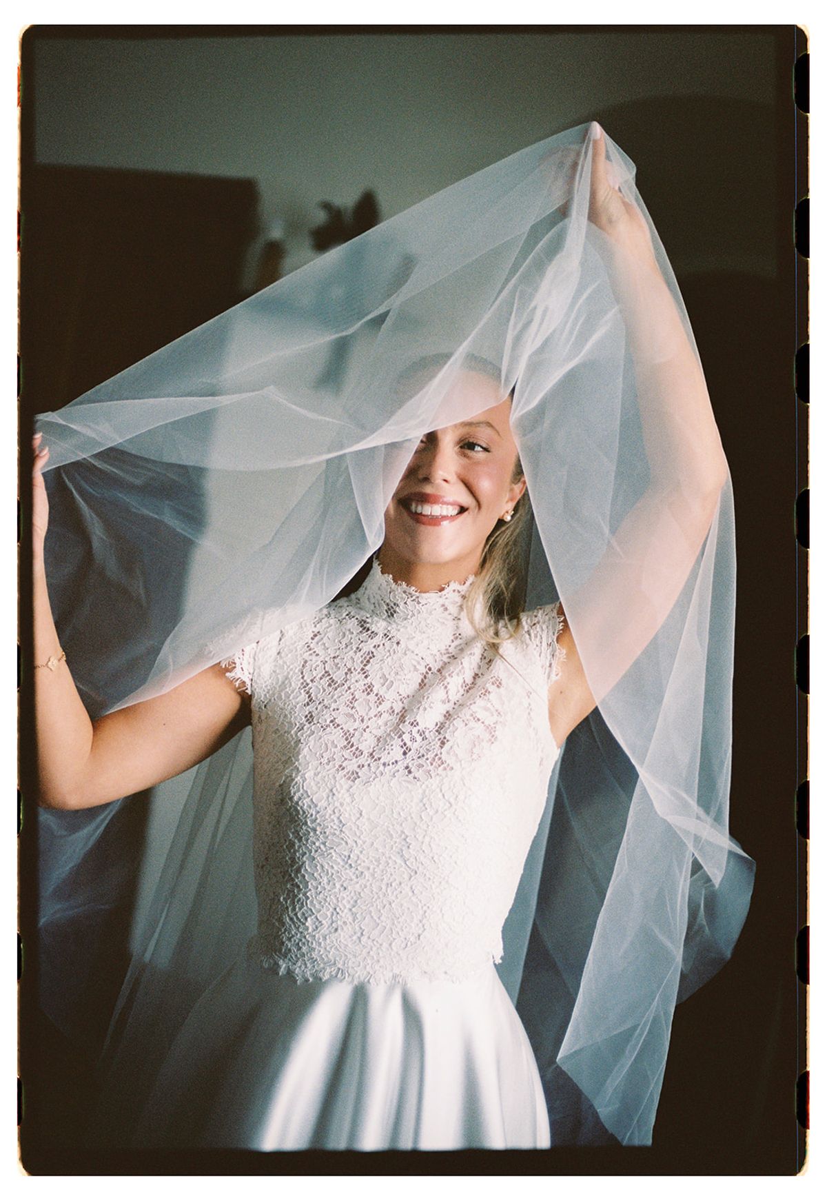 The bride in a lace high-neck dress smiles as she lifts a long sheer veil above her head.