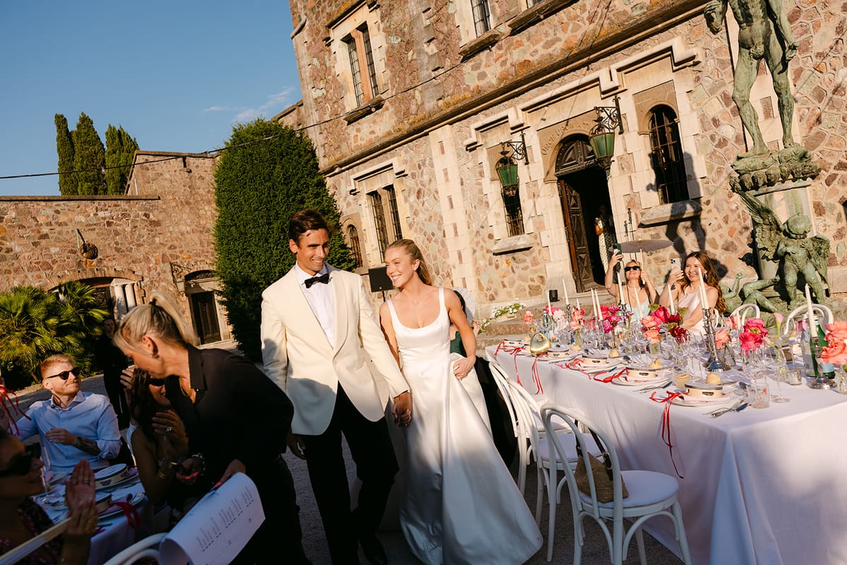 The couple walk beside a long table set with pink flowers and glasses, in front of a stone building, with guests around them.