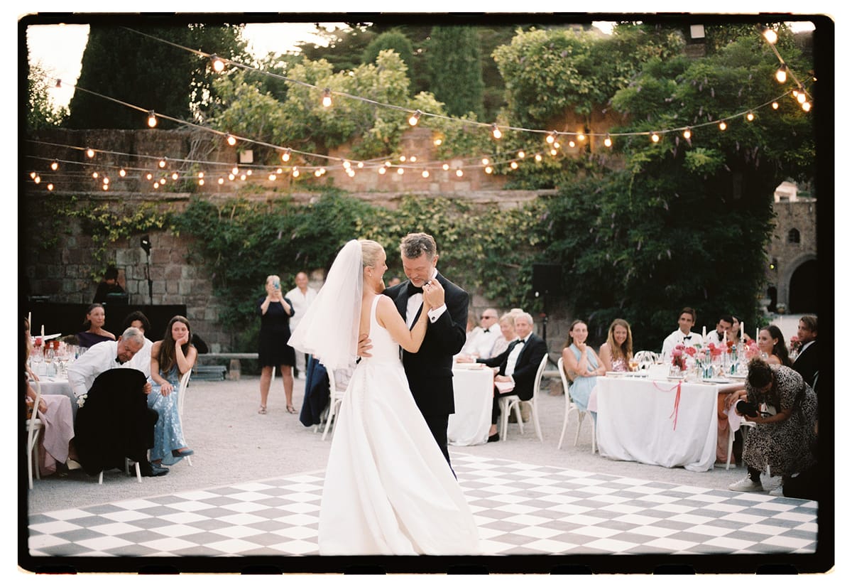 The couple dance together on a chequered floor in a courtyard while seated guests at round tables look on beneath string lights.