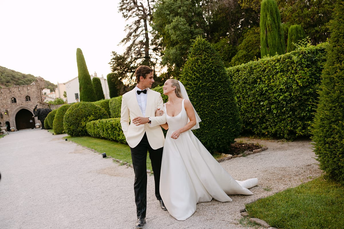 The groom in a white jacket and the bride in a white gown walk along a gravel path beside clipped hedges.