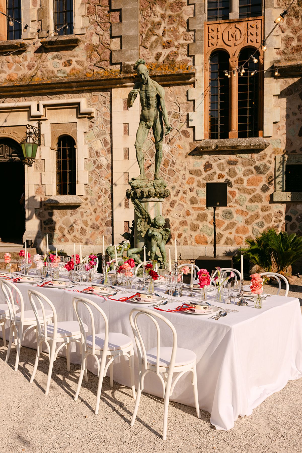 A long white-clothed table with bistro chairs and pink flowers stands on gravel in front of a stone building and statue.