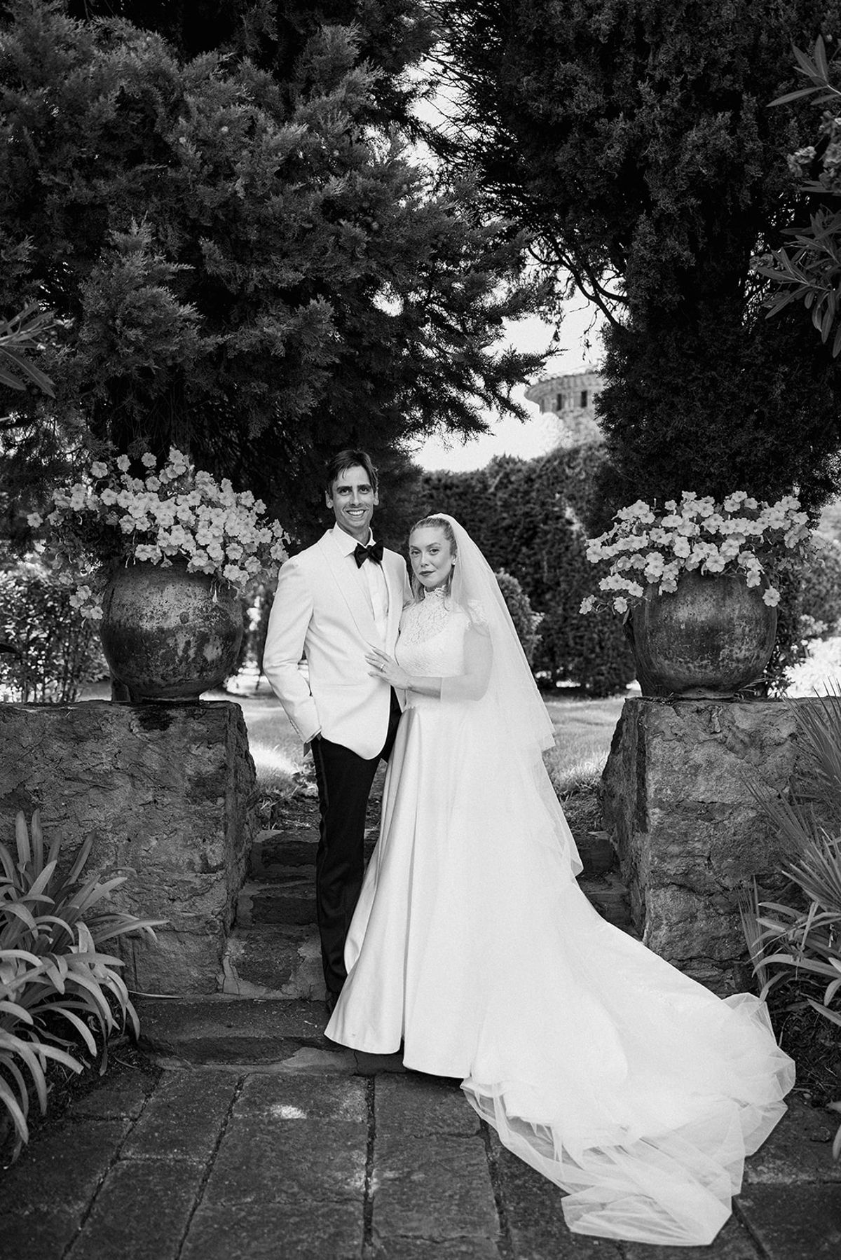 In black and white, the groom in a white jacket and the bride in a veil and long train stand together between flower pots.