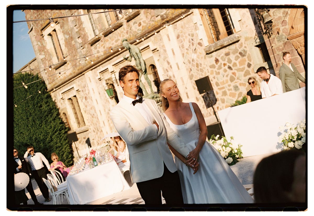 The groom in a white jacket and the bride in a white dress stand together in front of a stone chateau, with guests behind.