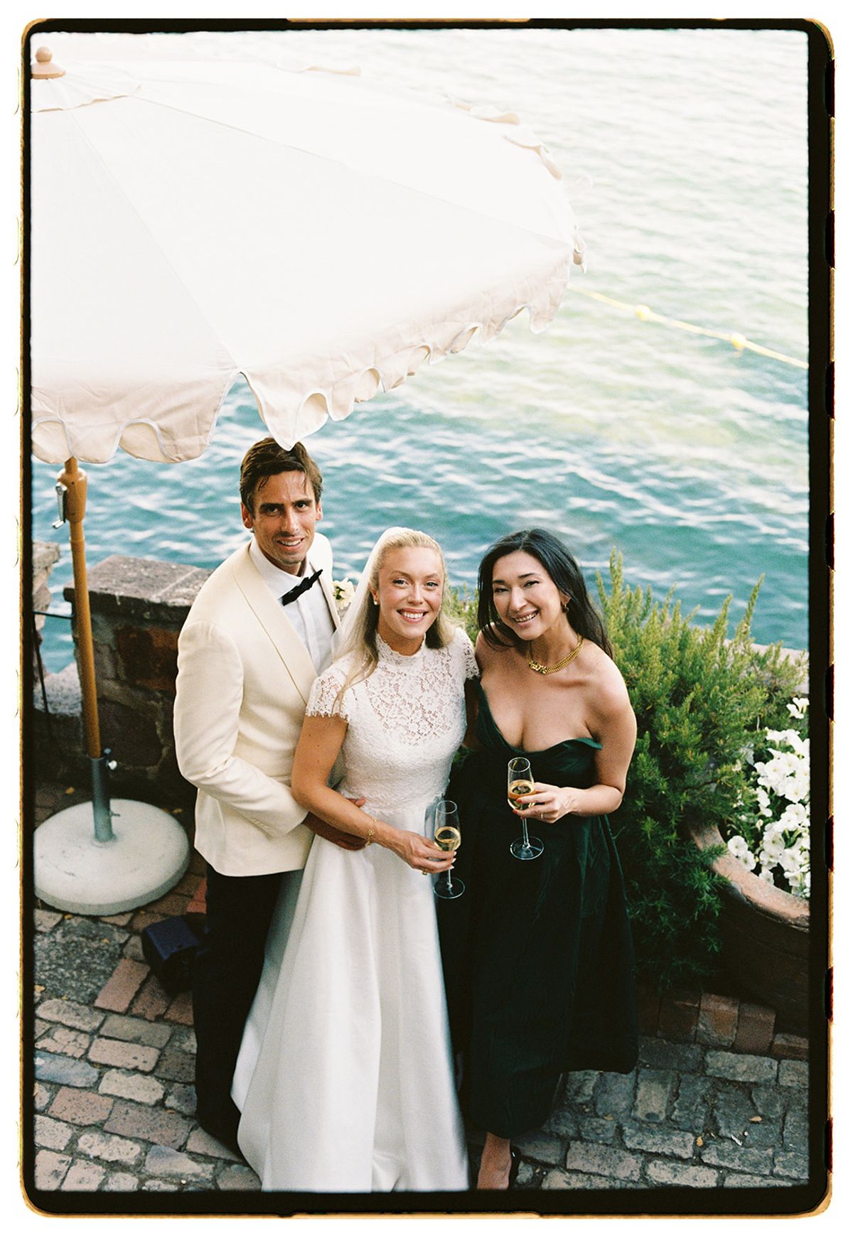 The couple and a guest in a black dress smile with drinks on a terrace beneath a white parasol above the sea.