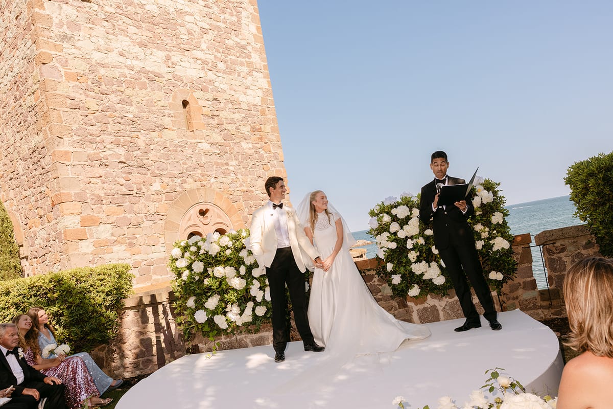 The groom, the bride in a long veil and an officiant stand on a white platform beside flowers, a stone tower and the sea.