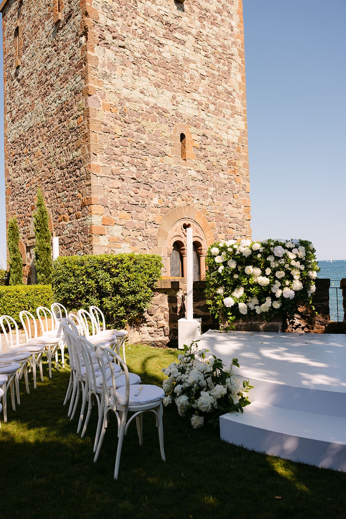 A tall stone tower rises behind rows of white chairs and a white platform decorated with white flowers on a lawn.
