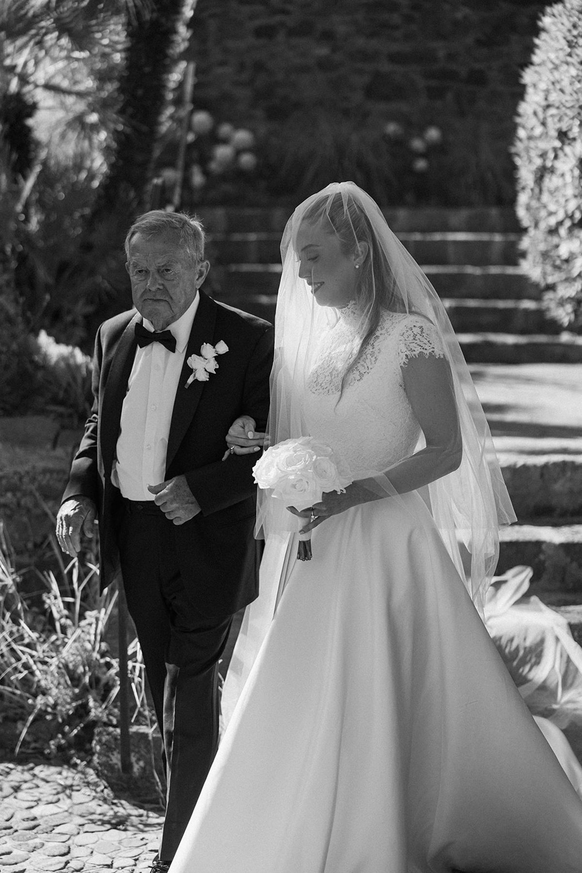 In black and white, the bride holding a bouquet walks beside a man in a dark suit down garden steps.