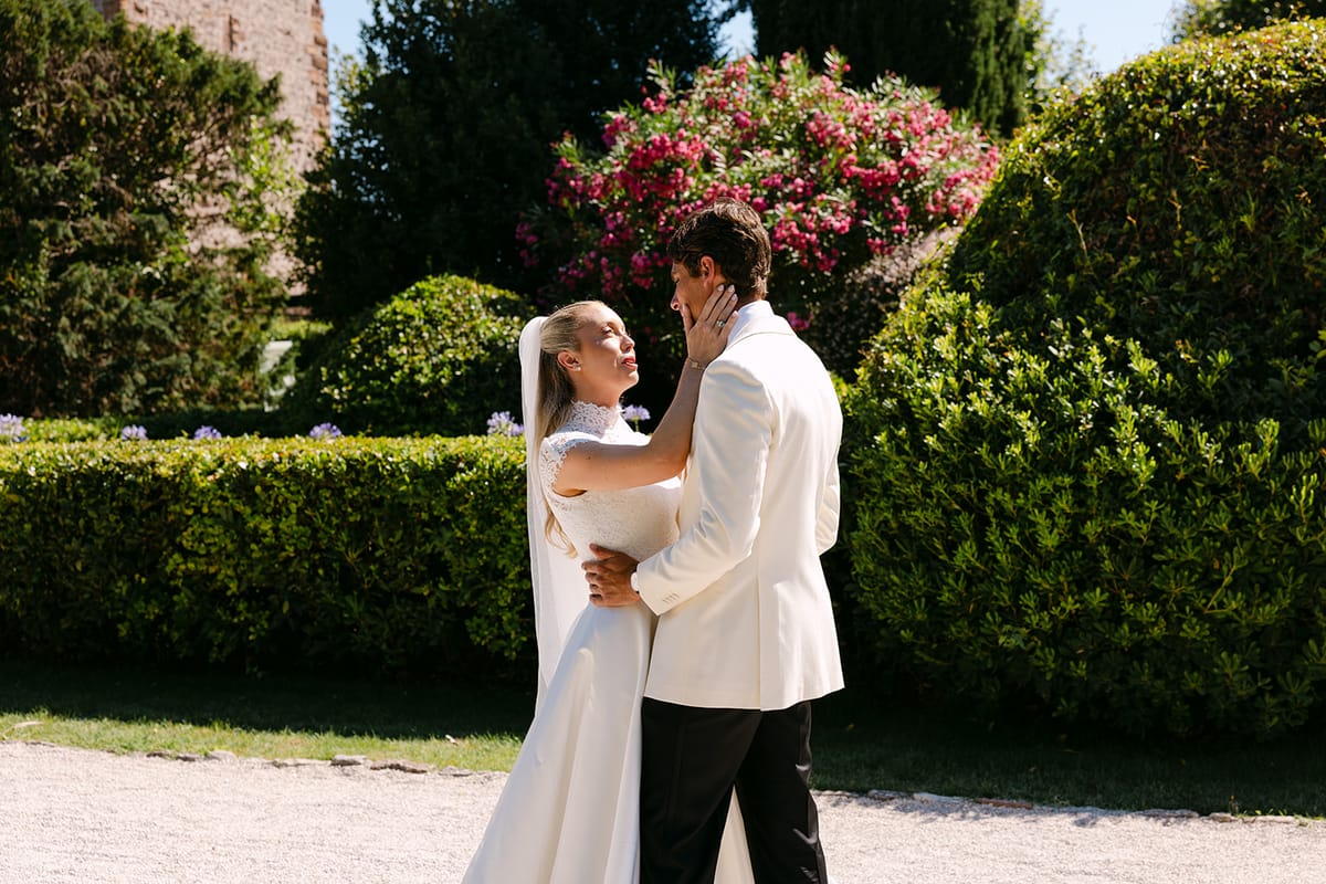 The bride and groom in white embrace on a gravel path in front of green hedges and pink flowering shrubs.