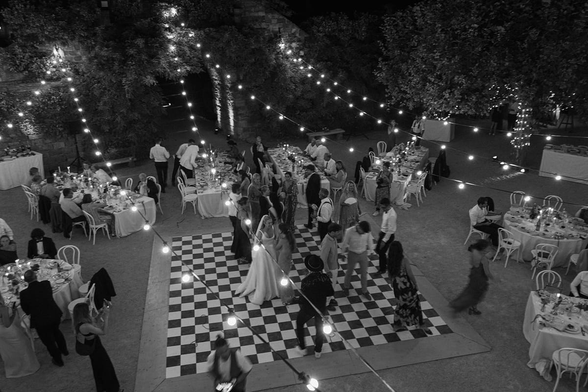 In black and white, seen from above, guests dance on a checkerboard floor beneath strings of lights, with dinner tables around.