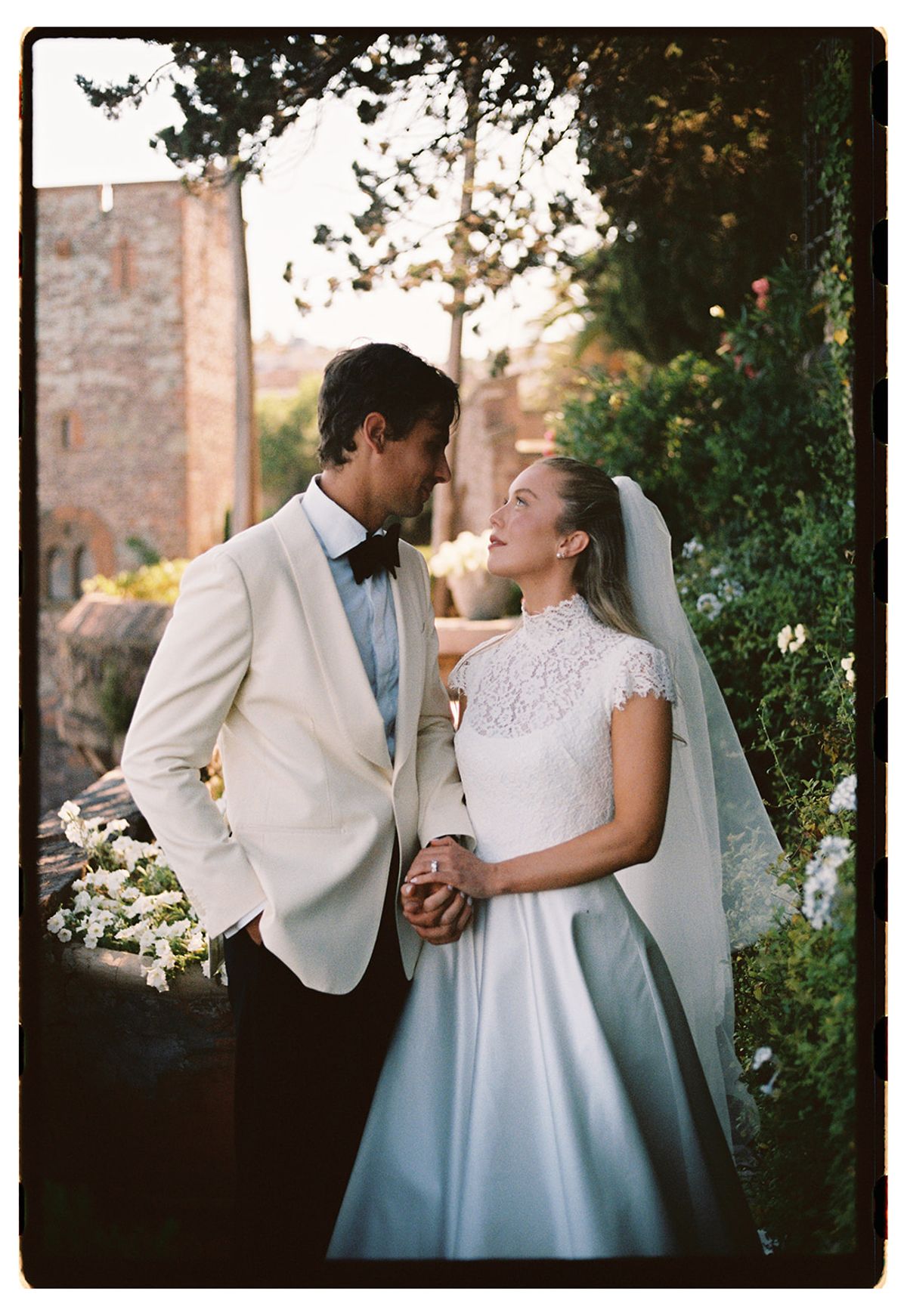 The groom in a white dinner jacket and the bride in a veil smile at each other, holding hands among greenery.