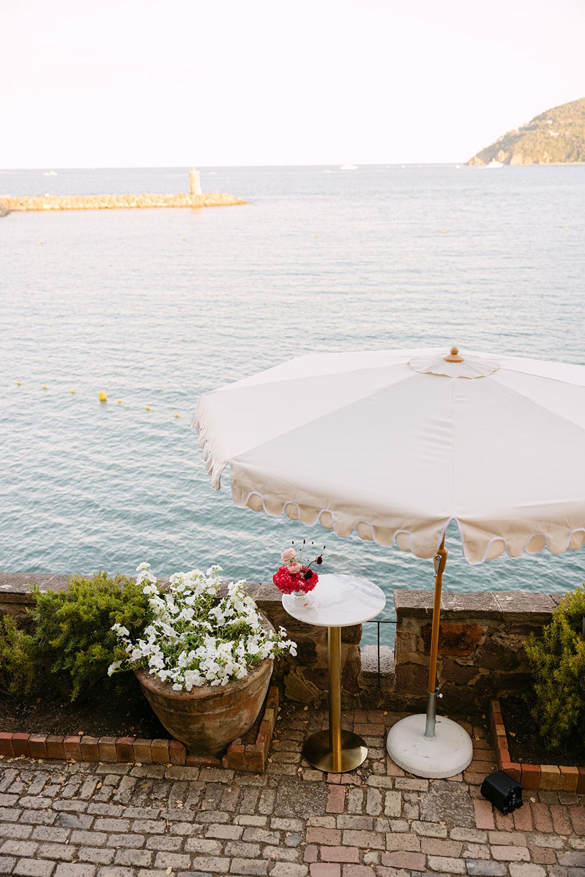 A cream parasol, a small table and pots of white flowers stand on a stone terrace overlooking calm sea.