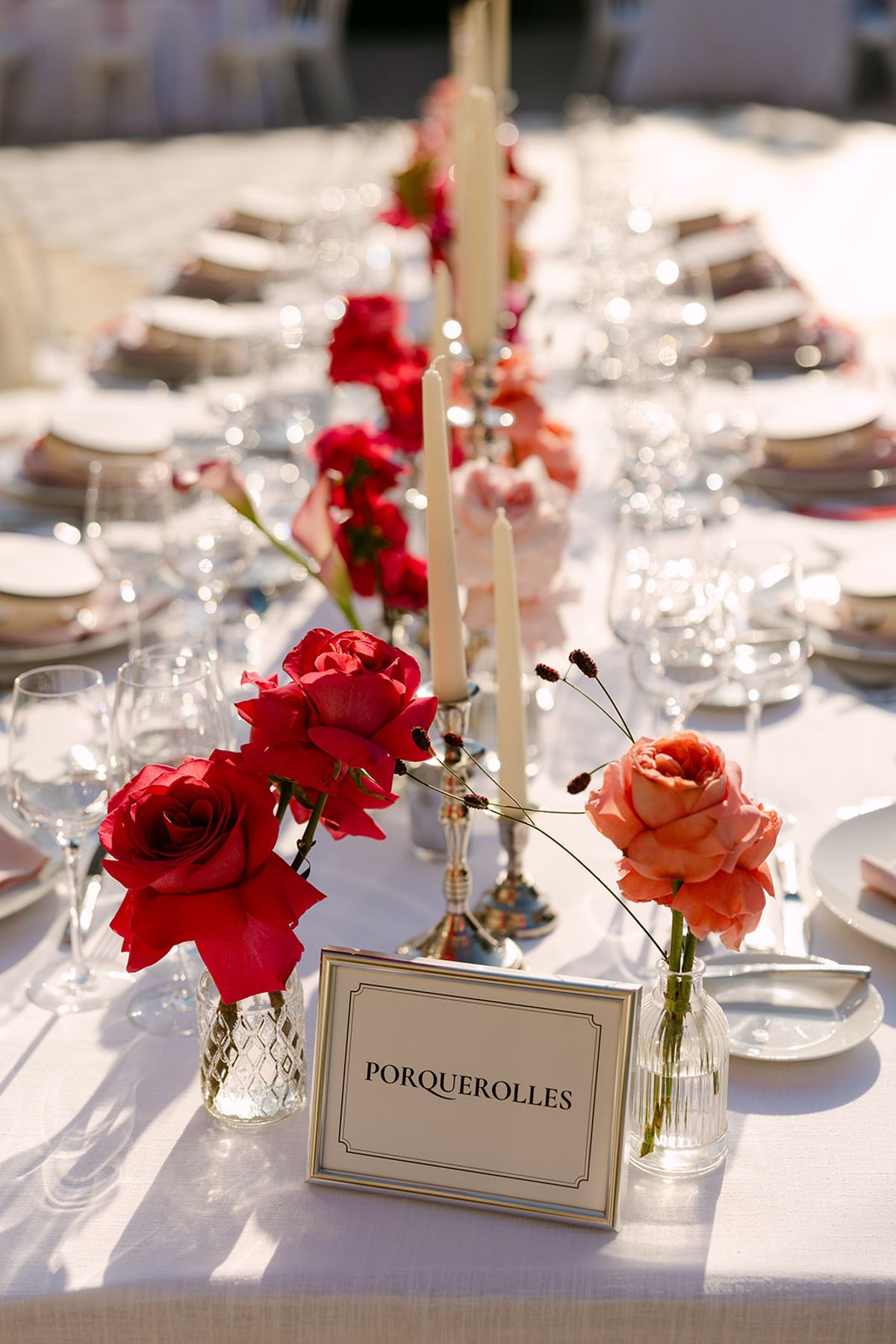 Red and coral roses in small vases stand beside tall candles and a framed table name card on a white tablecloth.