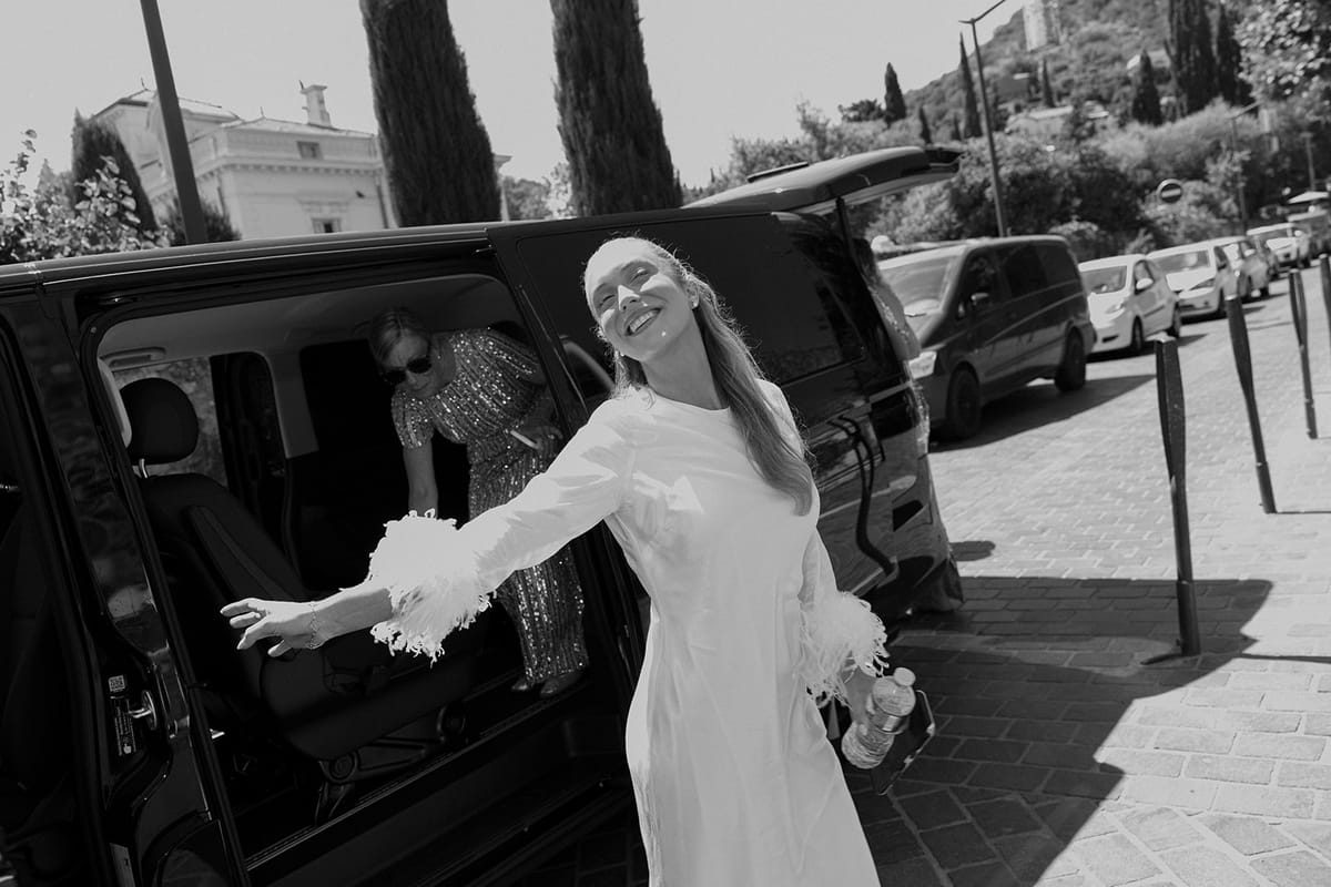 In black and white, the bride with feathered sleeves stretches out her arms beside an open van door on a street.