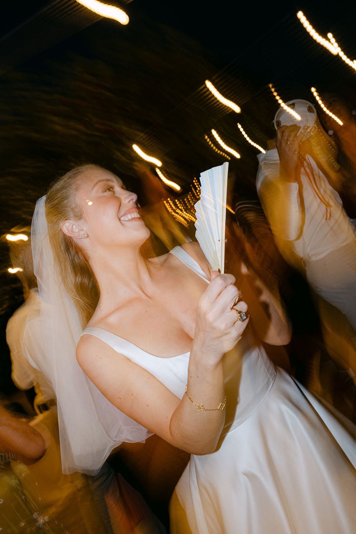 The bride in a veil laughs while holding a fan, with motion blur and streaks of light around her.