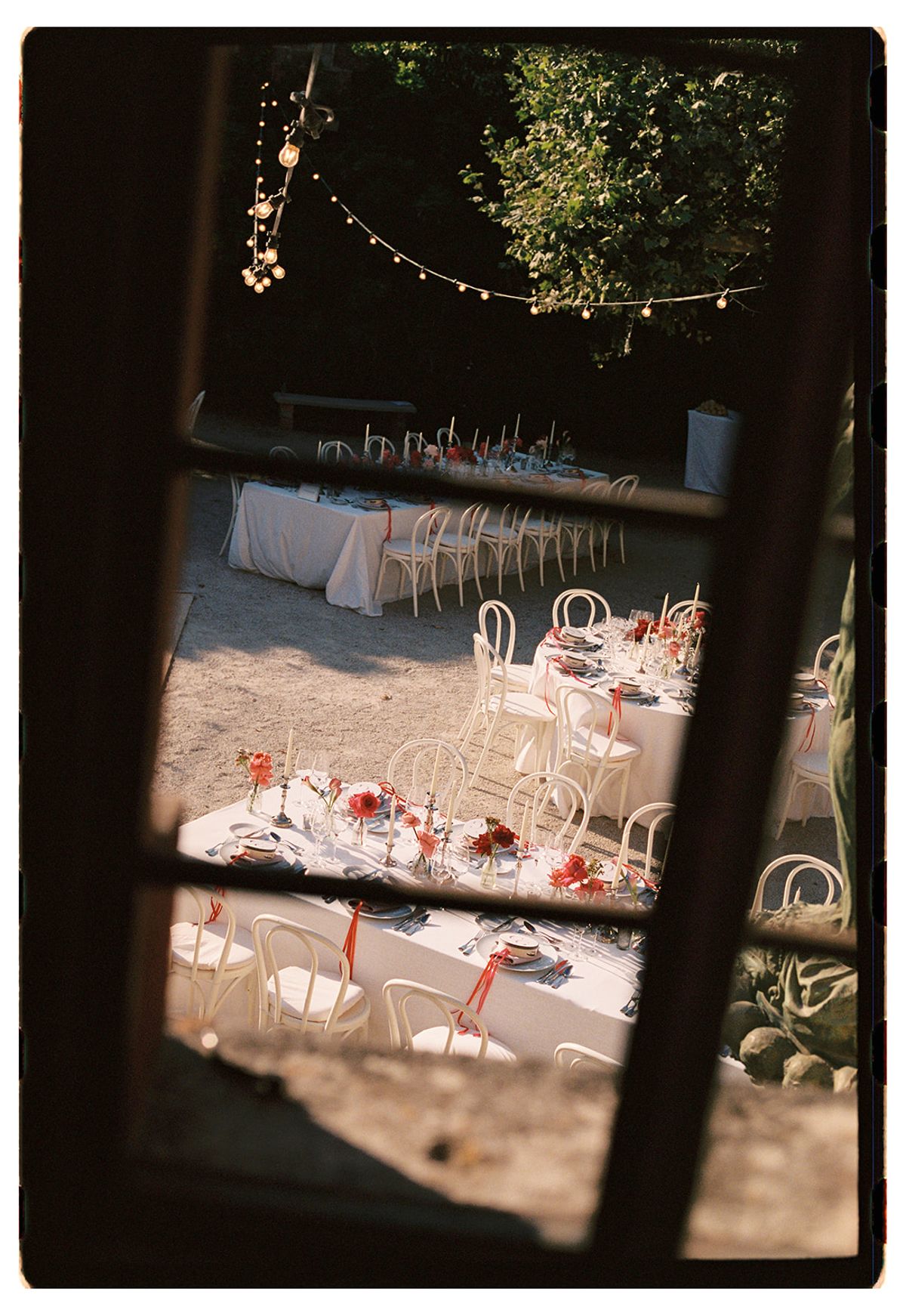 Through a dark window frame, set dinner tables and white chairs stand in a courtyard beneath strings of lights.