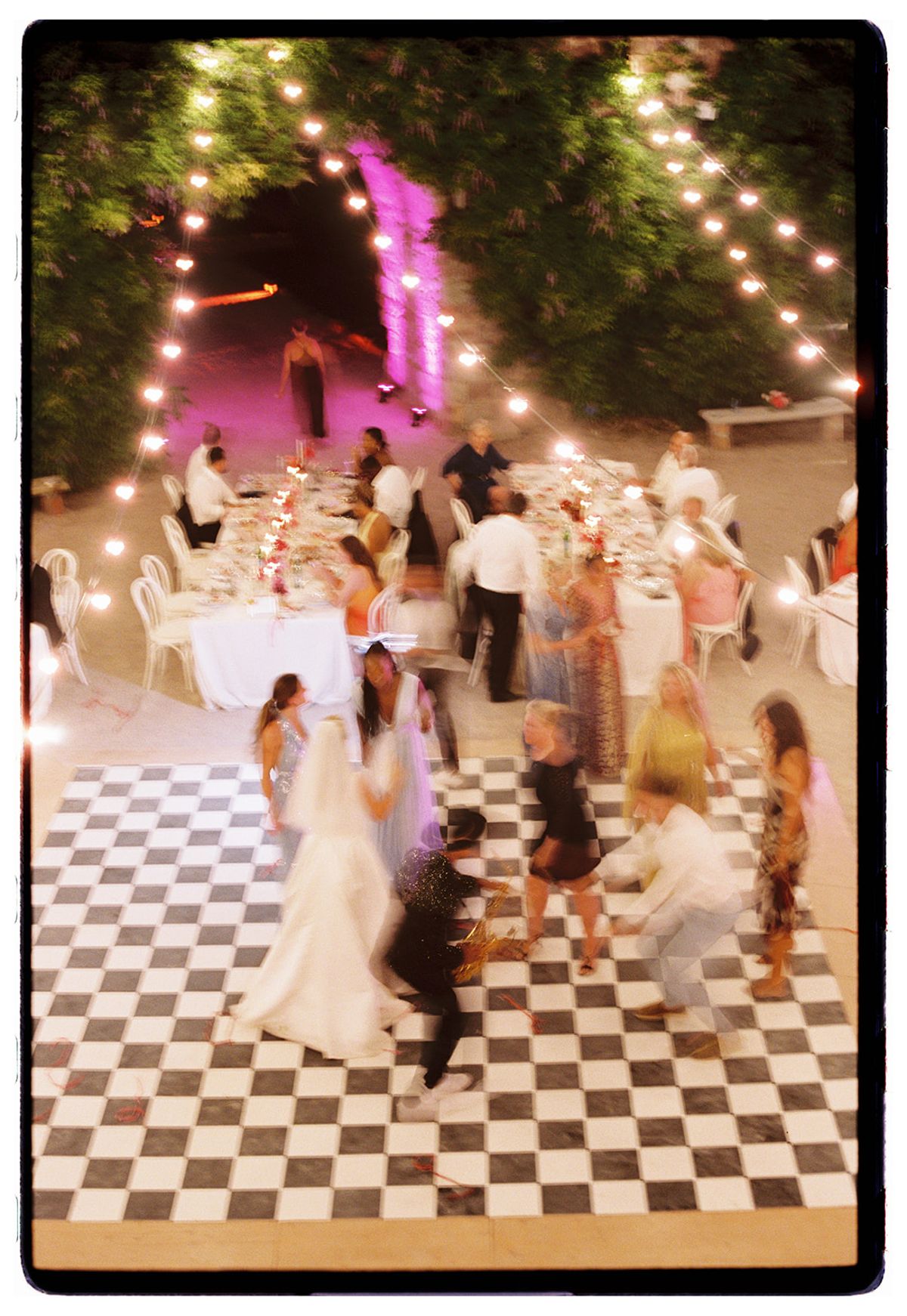 Blurred guests dance on a black and white chequered floor beneath string lights, with dining tables set up behind them.