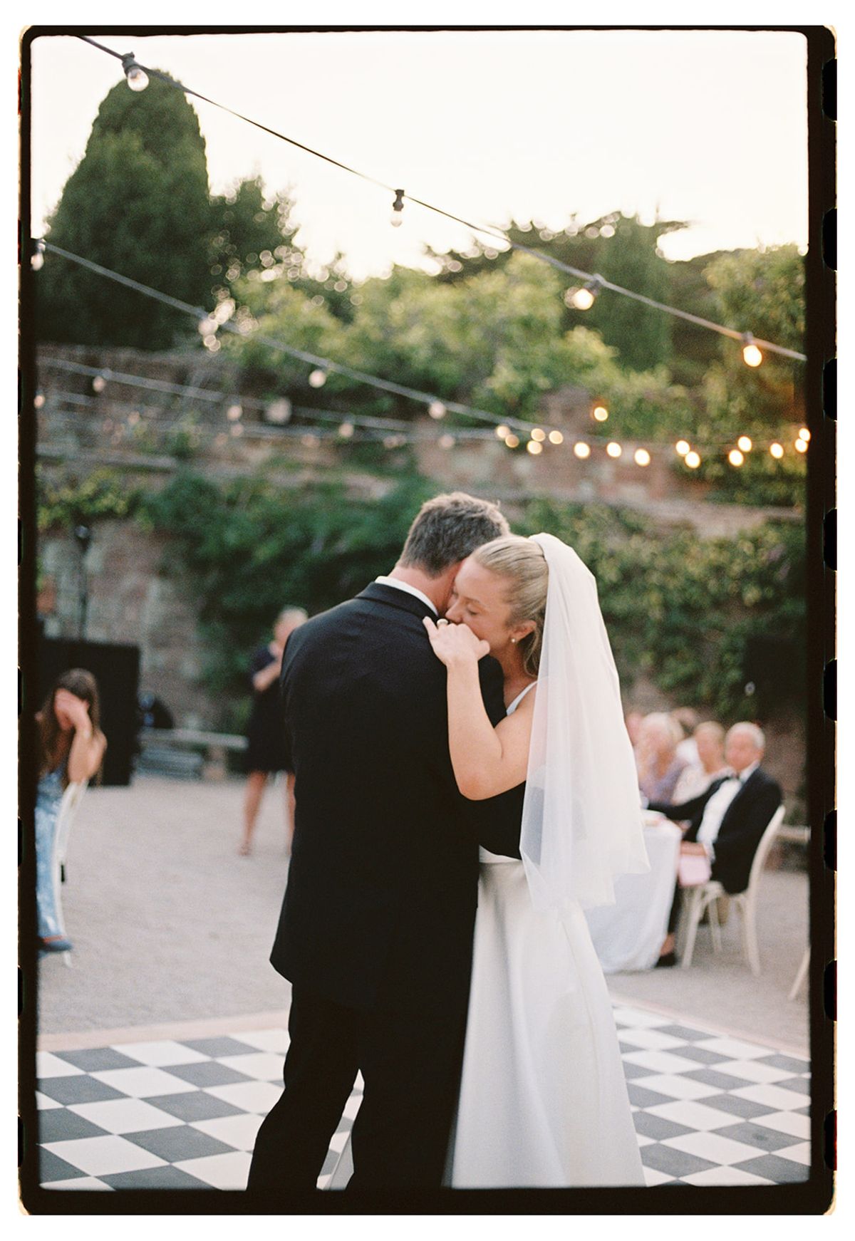 The couple embrace on a chequered dance floor beneath strings of lights, with guests seated in the background.