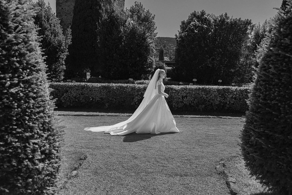 In black and white, the bride in a long gown with a train walks across a lawn between tall clipped conifers.