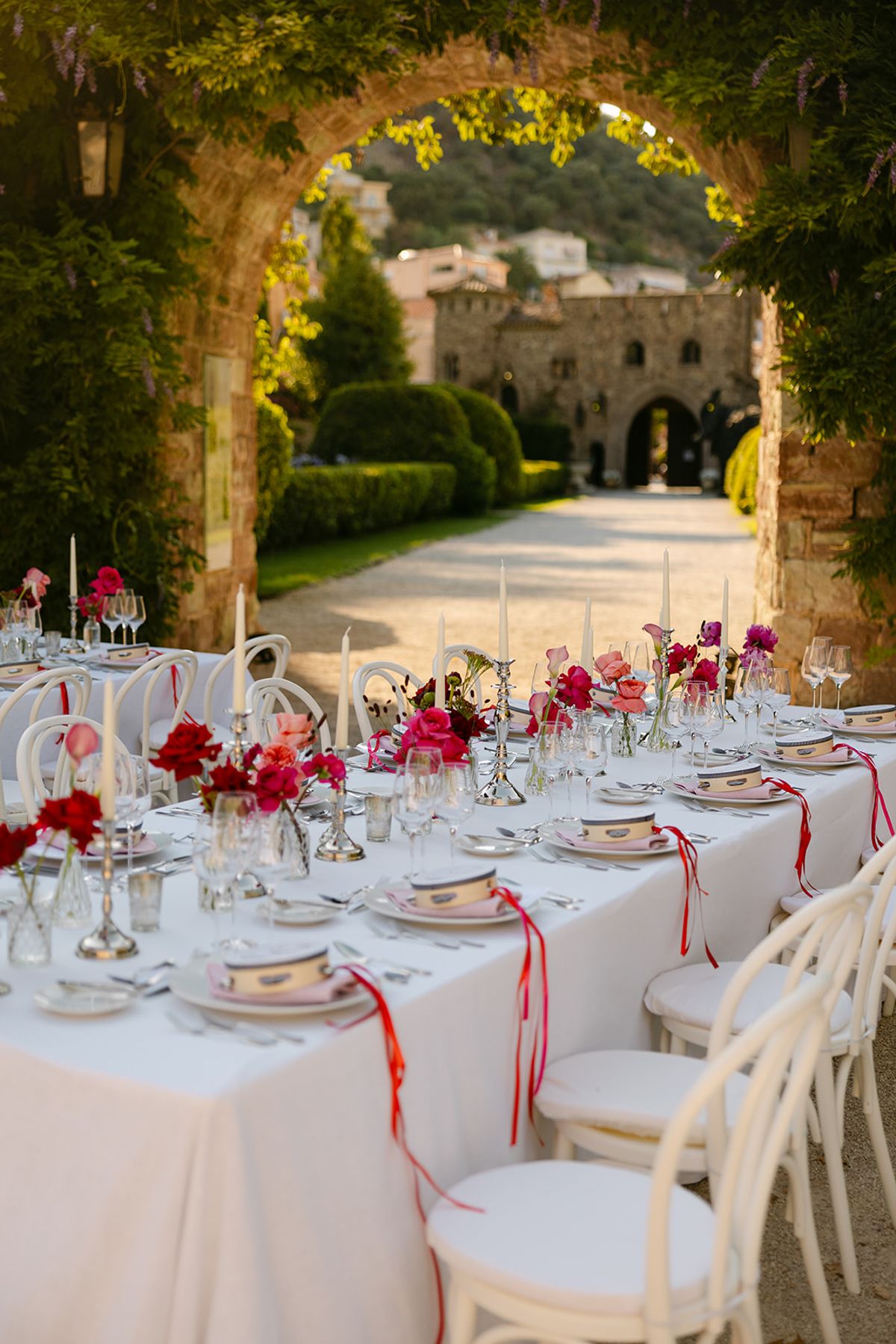 A long white-clothed table set with candles, red flowers and red ribbons stretches beneath a leafy stone archway.