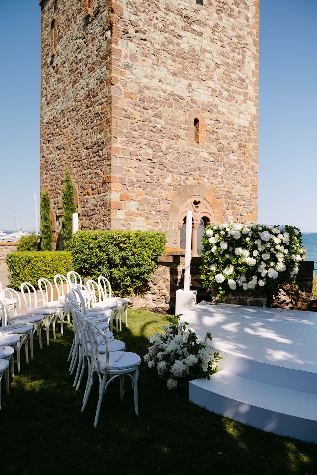 A tall stone tower stands behind white chairs on a lawn and a white platform dressed with white flowers.