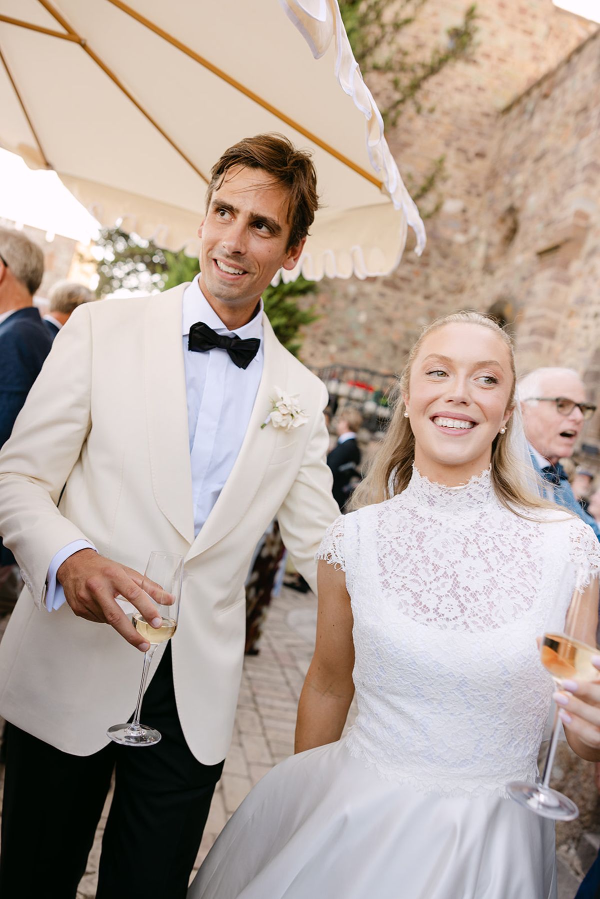 The groom in a white jacket and bow tie and the bride in a lace-collared dress smile, holding champagne glasses outdoors.