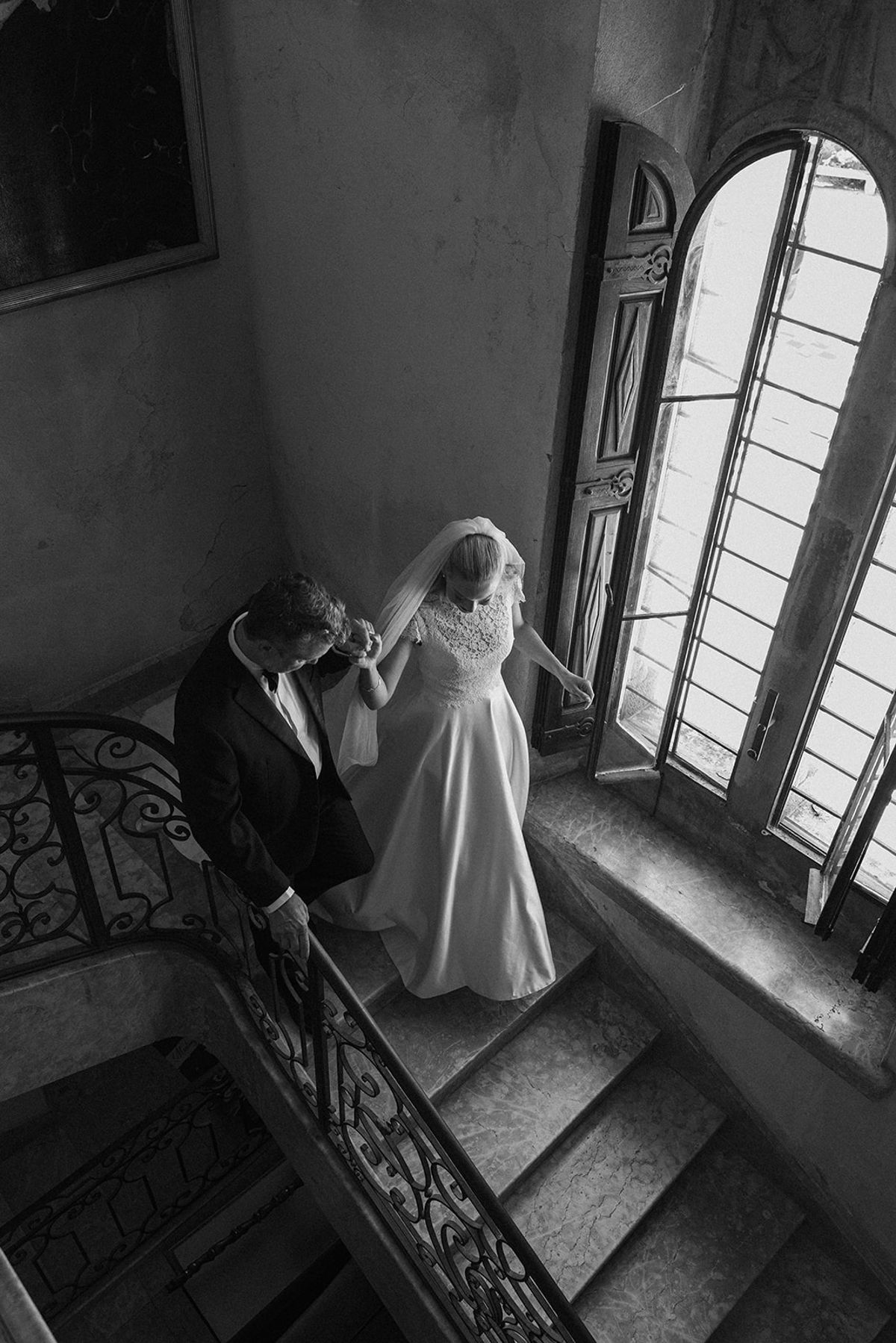 In black and white, the bride in a veil walks down a staircase beside a tall arched window, with a second person nearby.