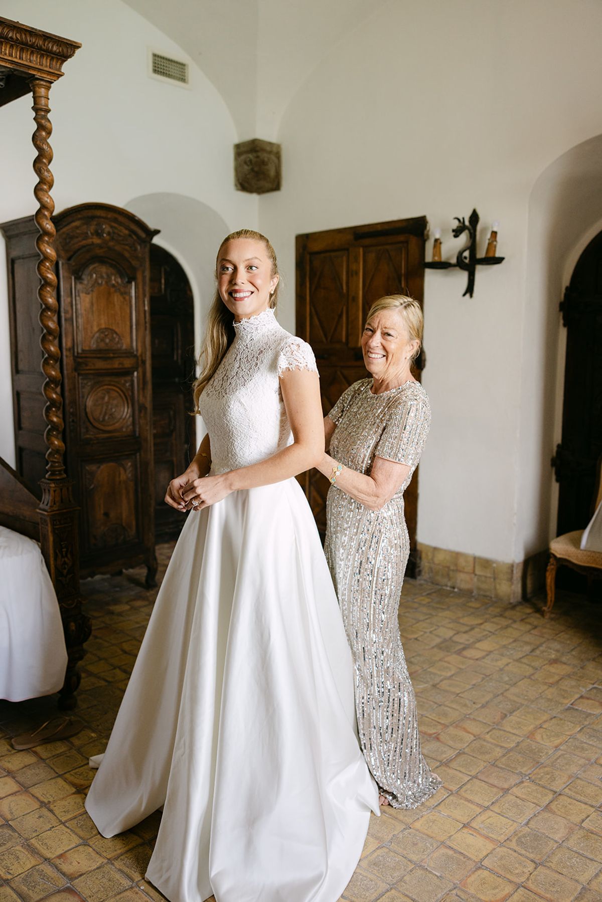The bride in a high-necked white gown stands in a stone-walled room as a woman in a sequinned dress fastens it behind her.