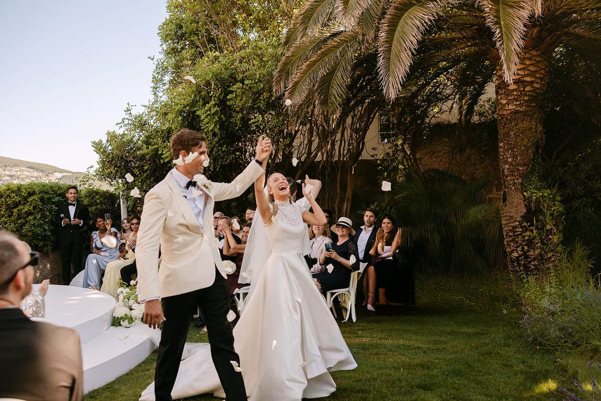 The groom raises an arm and the bride looks up as they leave the ceremony in a garden with palms, guests behind.