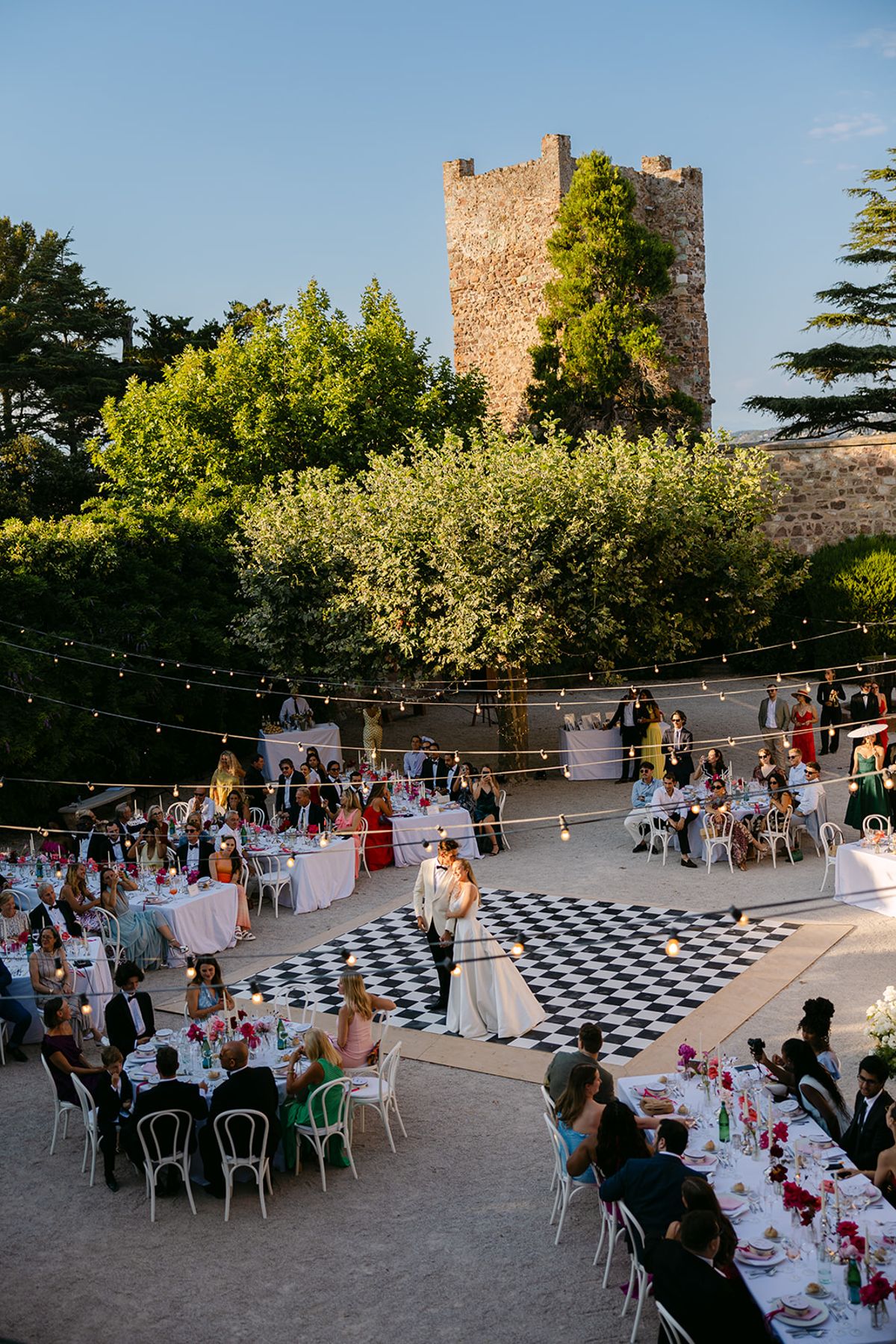 Seen from above, the couple dance on a black and white checkered floor with guests at tables around them below a stone tower.
