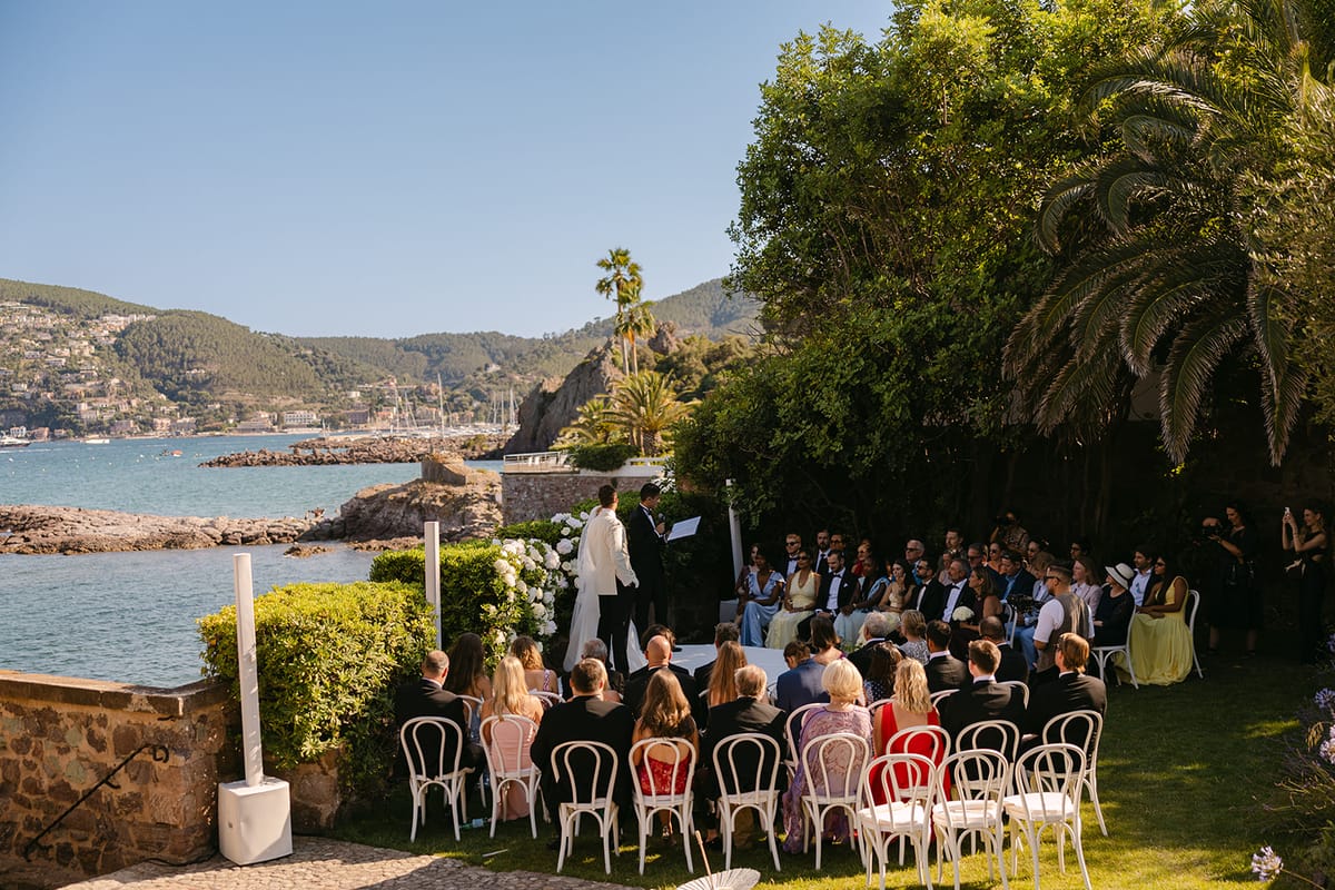 Rows of guests sit on white chairs in a garden terrace overlooking a bay, with hills in the distance under a blue sky.