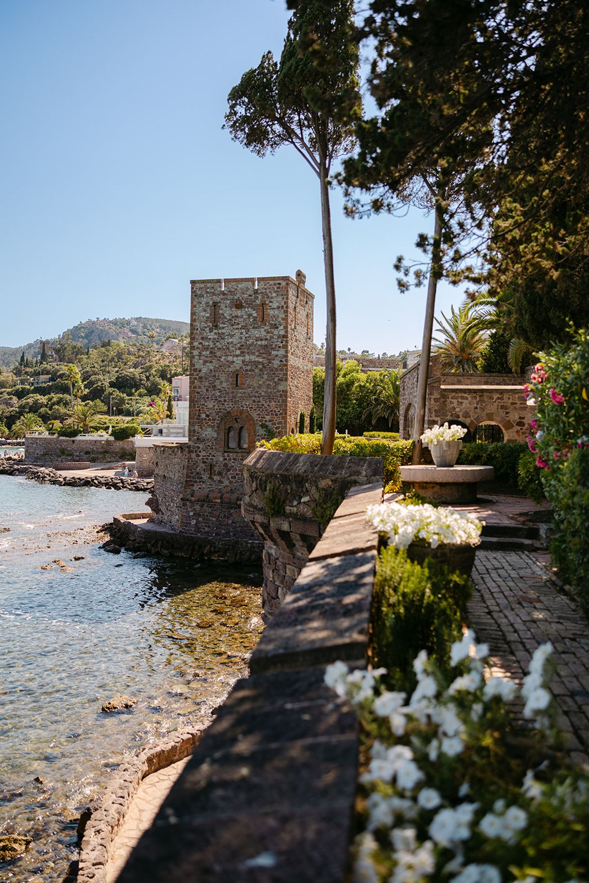 A stone tower stands beside the sea on a terrace wall lined with white flowers under a clear blue sky.