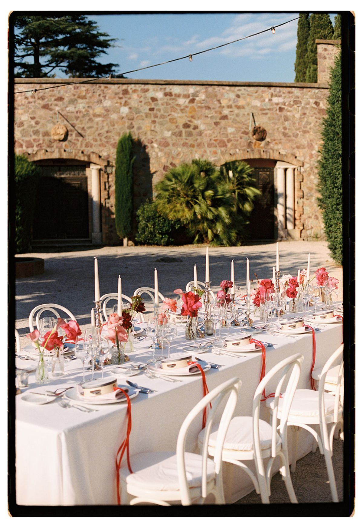 A long table with white cloth, red flowers, tall candles and white chairs stands in a stone courtyard beneath string lights.