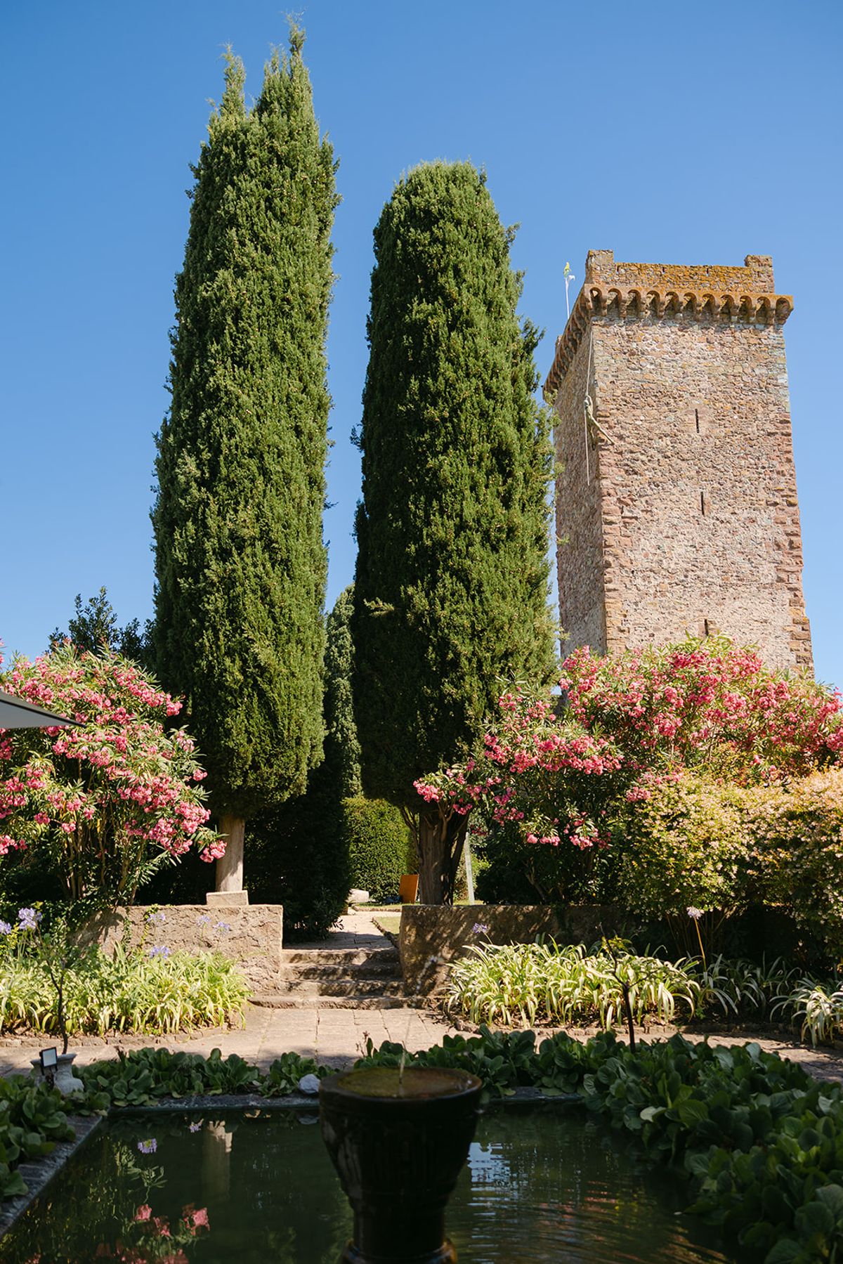 Two tall cypress trees and a stone tower rise above flowering shrubs and a pond under a clear blue sky.