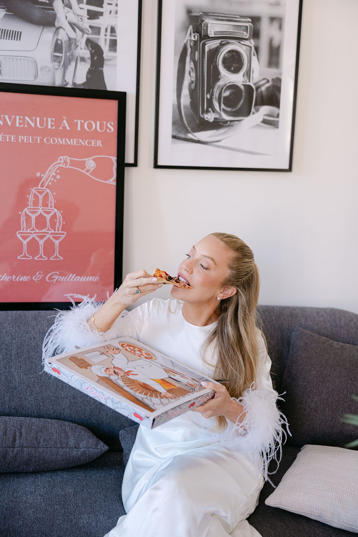 The bride, in a white dress with feathered sleeves, sits on a grey sofa eating a slice of pizza from a box.