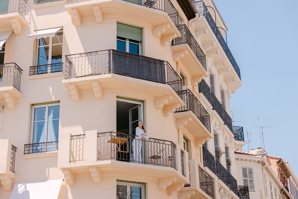 A cream coloured building with wrought iron balconies stands against a bright blue sky, with one person standing on a balcony.