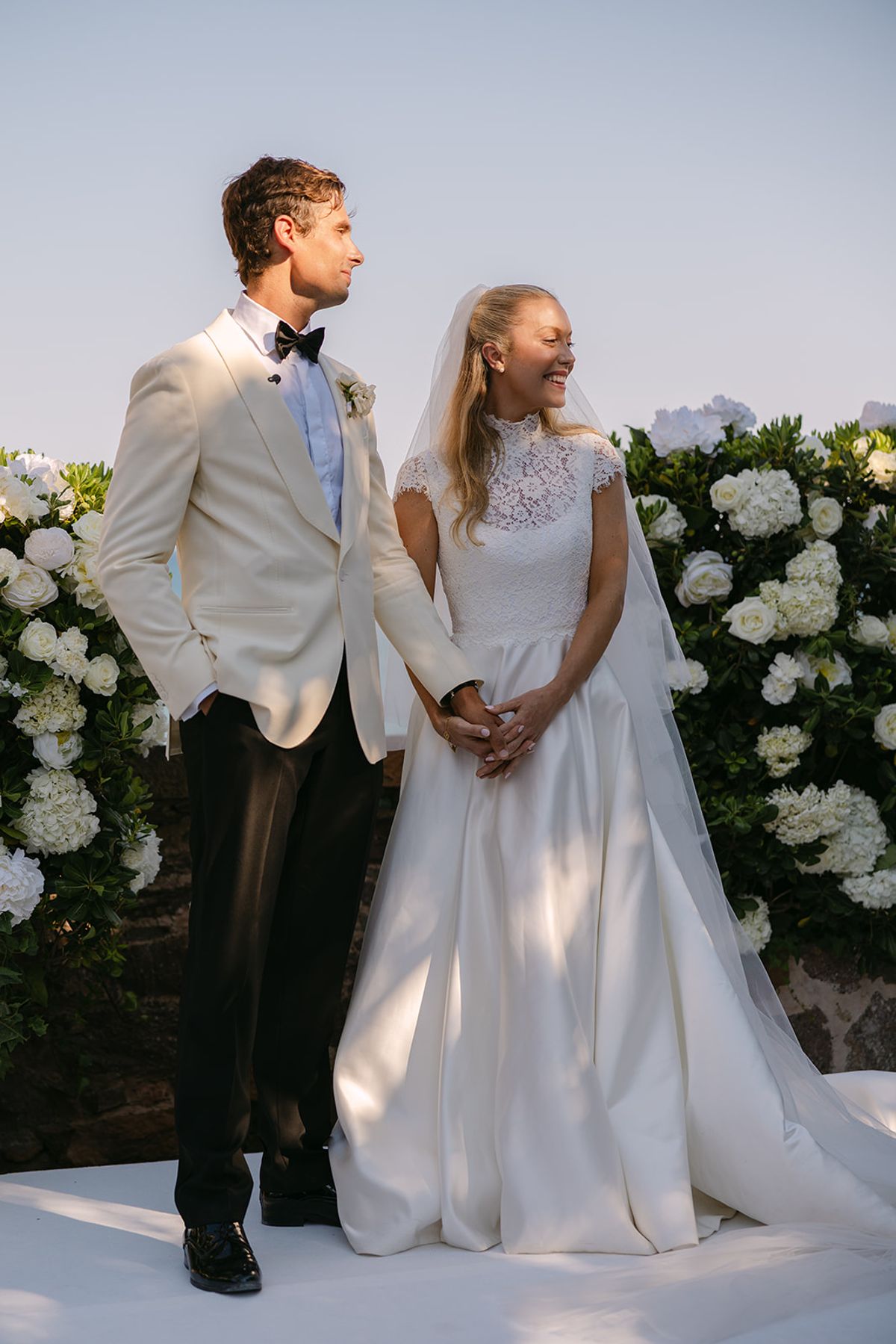 The groom in a white dinner jacket and the bride in a full gown hold hands on a white platform in front of banks of white hydrangeas.