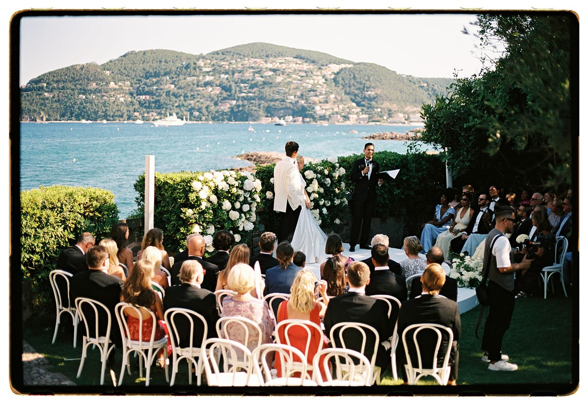 Guests sit on white bistro chairs facing the couple at a floral altar beside the sea, with a hilly coastline beyond.
