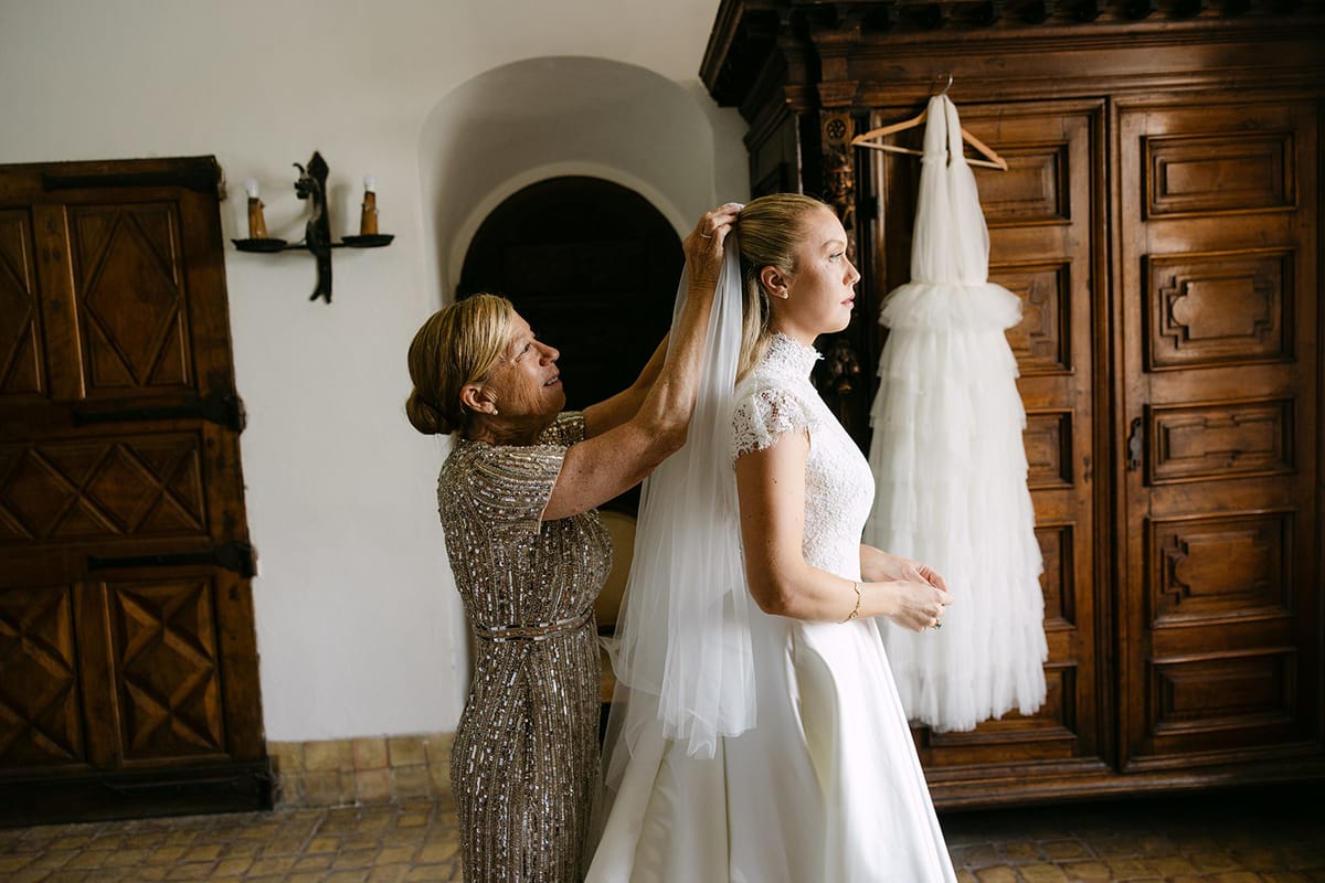 A woman in a sparkling dress adjusts the bride's veil in a room with a wooden wardrobe and a dress hanging beside it.