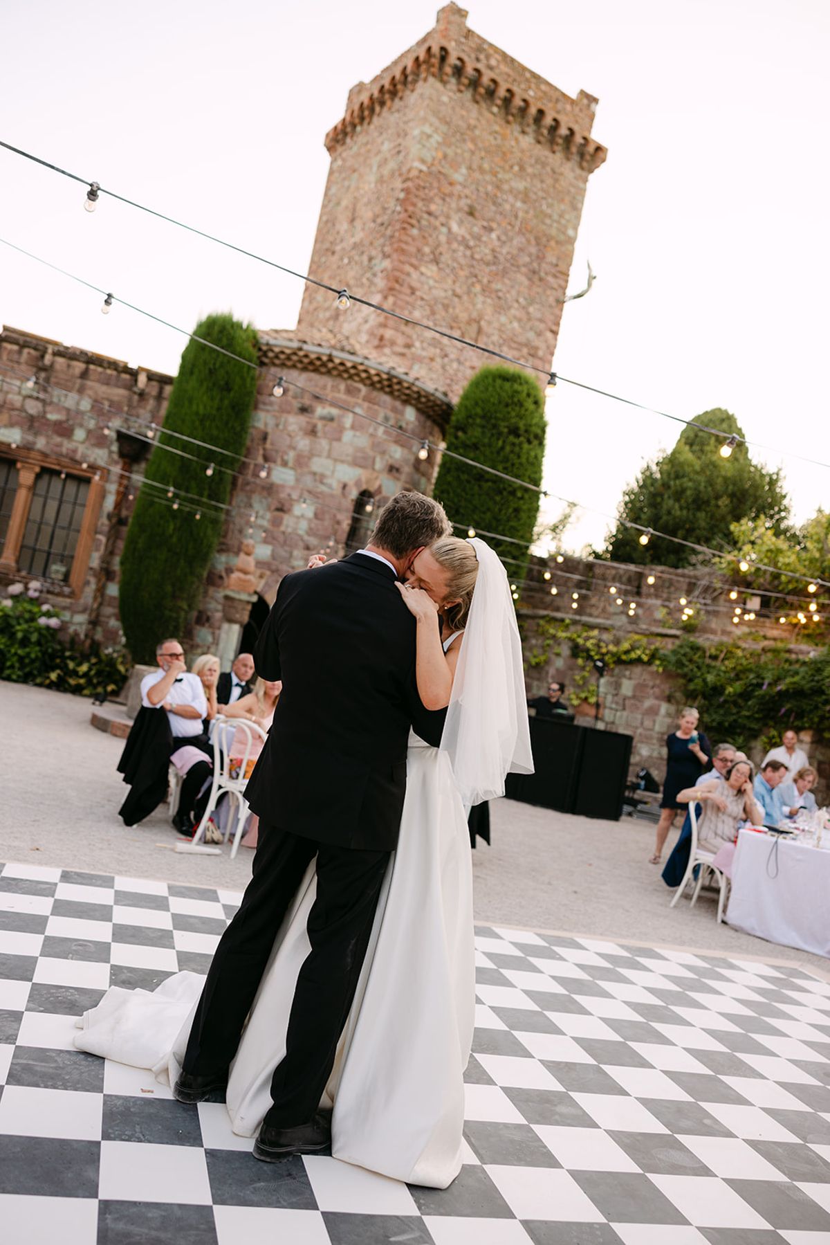 The couple share a kiss mid-dance on a chequered floor beneath a tall stone tower and clipped cypress trees.