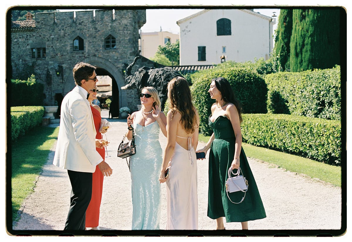 The groom in a white jacket stands with several women in light blue, cream and green dresses on a gravel path in front of a stone archway.